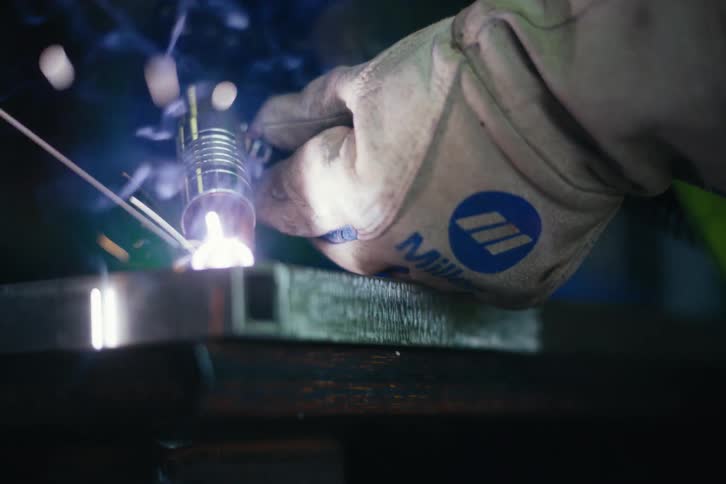 Close-up slow motion shot of a welder in a protective helmet joining square metal tubing with a welding torch, creating bright sparks and smoke
