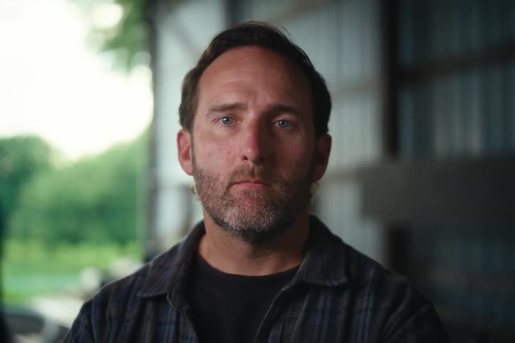 Close-up portrait of a serious middle-aged man with a beard looking up at the camera inside a rustic barn