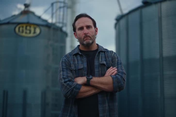 Worried farmer with arms crossed looks up at the camera with a serious expression in front of two grain silos