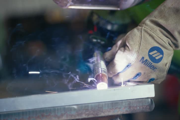 A close-up cinemagraph of a welder's gloved hand using a torch to fuse metal, creating bright sparks and smoke