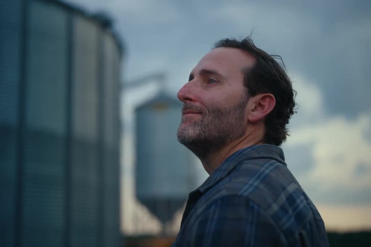 Side profile of a contemplative farmer looking up with a slight smile, with large grain silos in the background at dusk