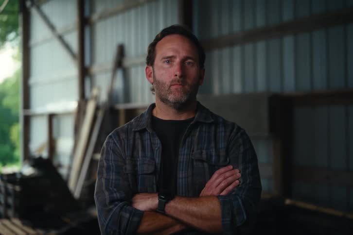 A rugged man with a beard crosses his arms and looks directly at the camera while standing inside a barn