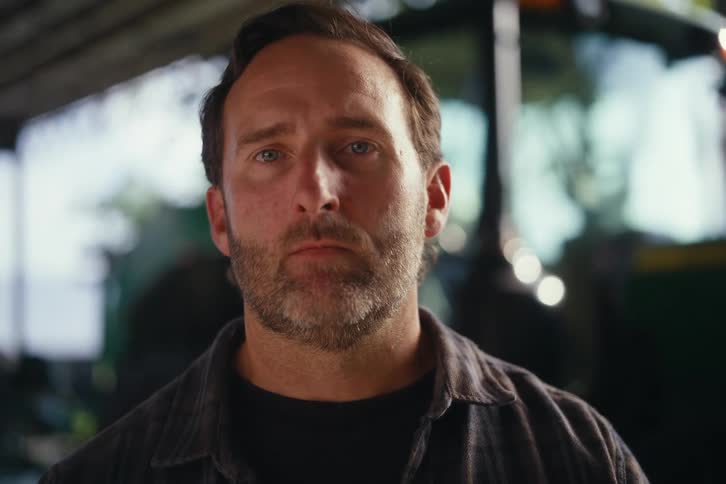 A close-up portrait of a serious farmer with a beard turning to look directly into the camera inside a barn