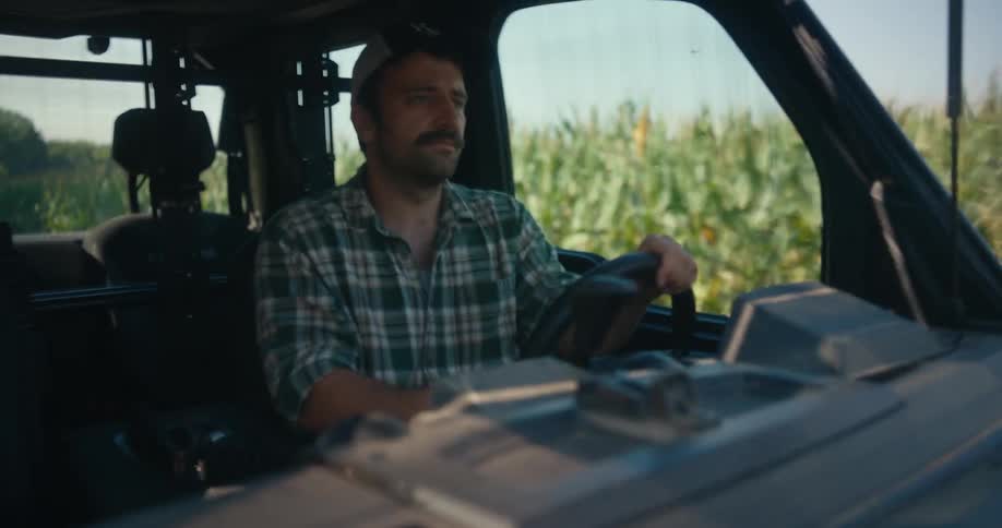 A mustached farmer in a plaid shirt and cap drives his utility vehicle alongside a vibrant green cornfield on a sunny day