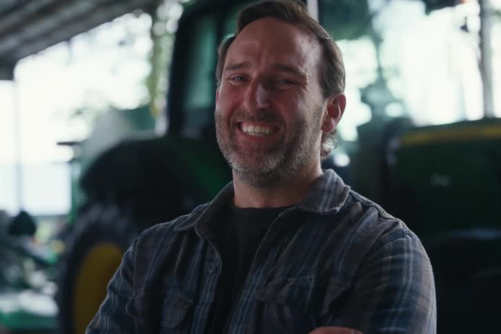 Portrait of a happy, bearded male farmer in a plaid shirt smiling at the camera, with a large green tractor in the background of a barn