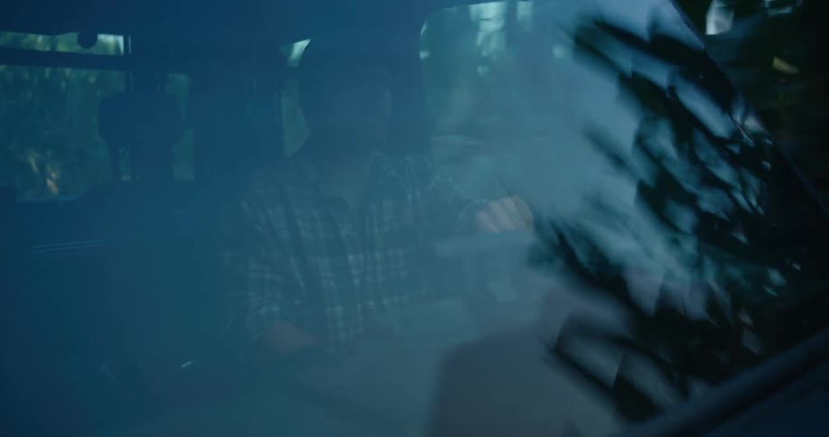 A man in a plaid shirt drives a vehicle at dusk, seen through the windshield with reflections of passing trees