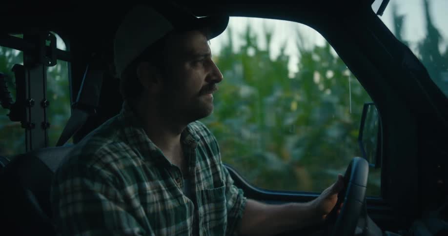 Close-up of a farmer driving his UTV alongside a cornfield, with the light fading from day to evening