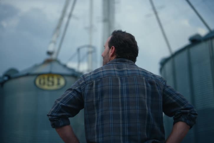 Back view of a farmer in a plaid shirt looking up proudly at his towering metal grain silos