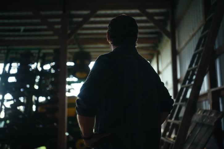 A thoughtful male farmer stands silhouetted in the doorway of a rustic barn, surveying his land at dawn