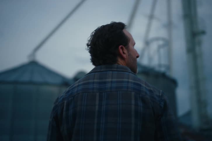 A thoughtful farmer in a plaid shirt surveys his grain silos during a moody, overcast evening