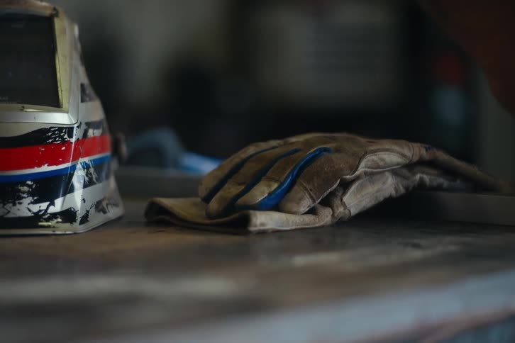 A close-up of a welding helmet and leather work gloves resting on a workbench in a dimly lit workshop
