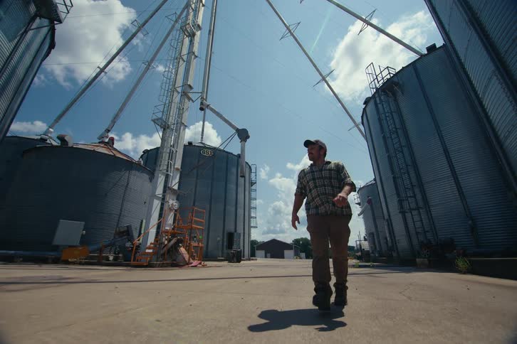 Low angle slow motion shot of a farmer walking with determination between towering grain silos under a bright blue sky