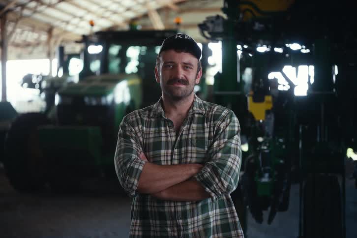 Confident male farmer with a mustache and arms crossed looks up and smiles while standing in front of tractors in a barn