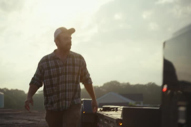 A farmer with a mustache wearing a plaid shirt and hat secures a flatbed trailer then walks to his truck in slow motion at sunrise