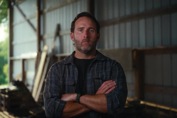 A resolute and serious-looking farmer crosses his arms as he looks directly into the camera inside a rustic barn
