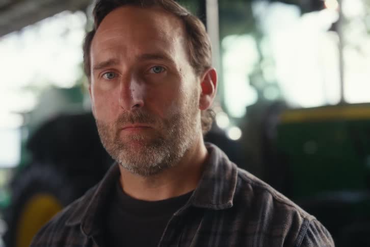 A rugged, serious farmer in a plaid shirt looks up with a determined gaze inside a barn