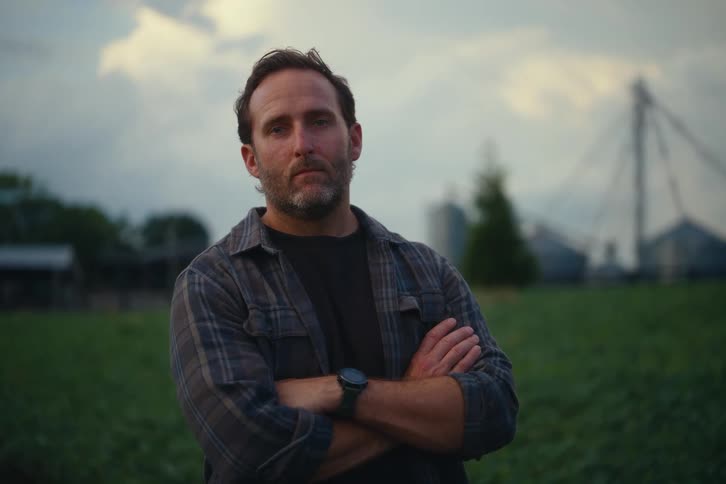 A proud farmer with his arms crossed stands in a green field at dusk and looks directly into the camera