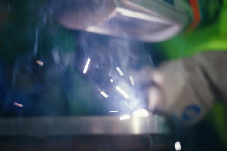 Close-up slow-motion shot of a welder's helmet tilting down to reveal a gloved hand using a torch to weld metal, creating bright sparks and smoke