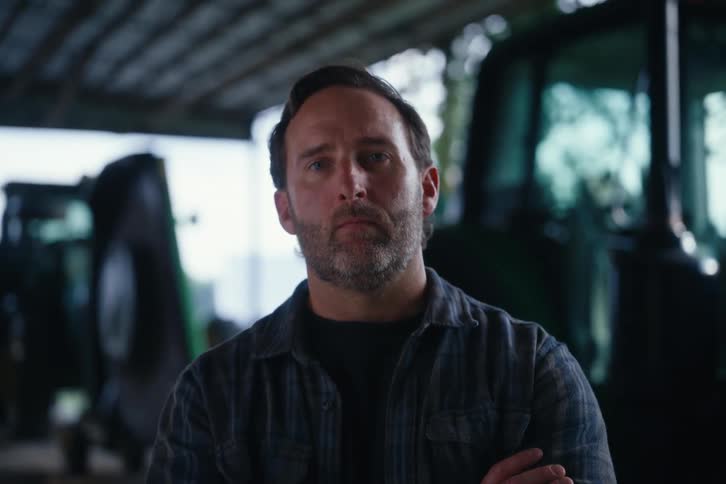 A serious, bearded farmer crosses his arms and looks at the camera while standing in front of a tractor in a barn
