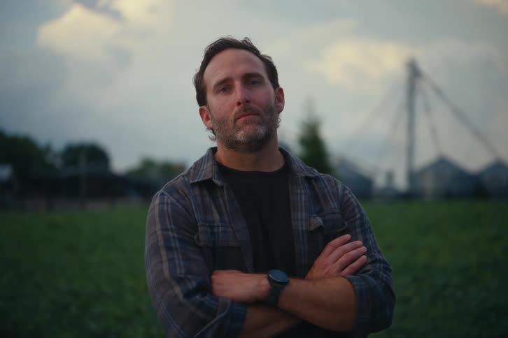 Portrait of a stoic farmer in a plaid shirt standing in a field with farm buildings in the background at twilight