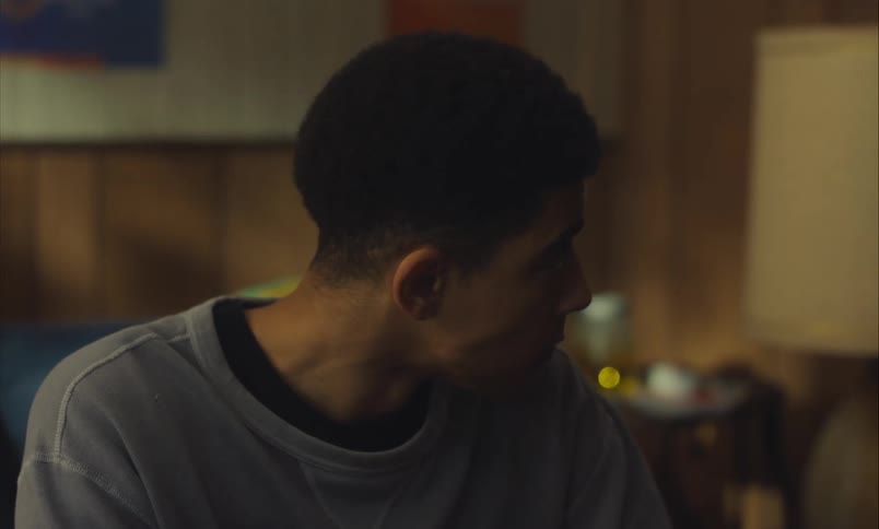 A thoughtful and serious young man looks down while sitting alone in a dimly lit, wood-paneled room