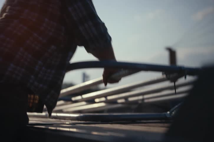 A farmer in a plaid shirt loads a metal gate onto a trailer in slow motion, silhouetted against the bright morning sun