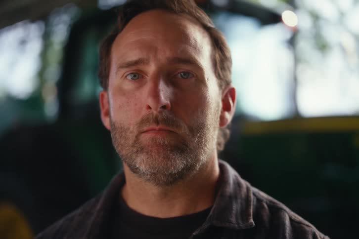 A serious, bearded man in a flannel shirt slowly looks up with an intense stare while standing in a barn