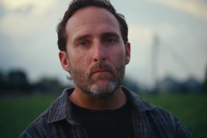 A dramatic rack focus reveals a close-up of a serious bearded man looking intently into the camera outdoors at dusk