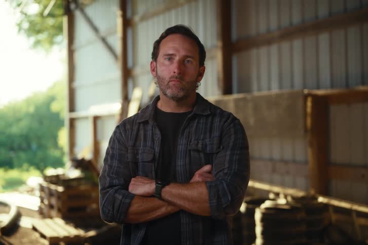 A contemplative man standing in a barn looks up with a serious and determined expression