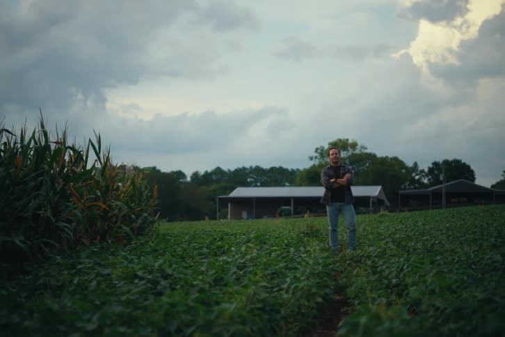 A proud farmer stands with arms crossed in the middle of his vast crop field, surveying his land under a cloudy evening sky