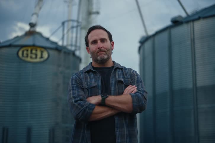 A serious farmer with his arms crossed stands proudly in front of two large grain silos