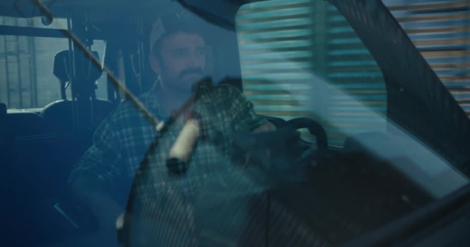 A mustachioed farmer in a plaid shirt and baseball cap drives a utility vehicle on his farm, with silos and fields reflecting in the windshield
