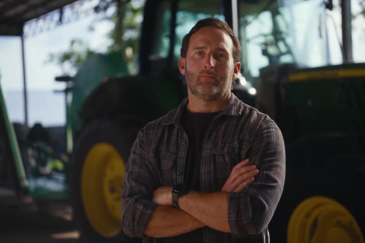 A serious farmer in a plaid shirt stands confidently in his barn with his arms crossed in front of his tractor