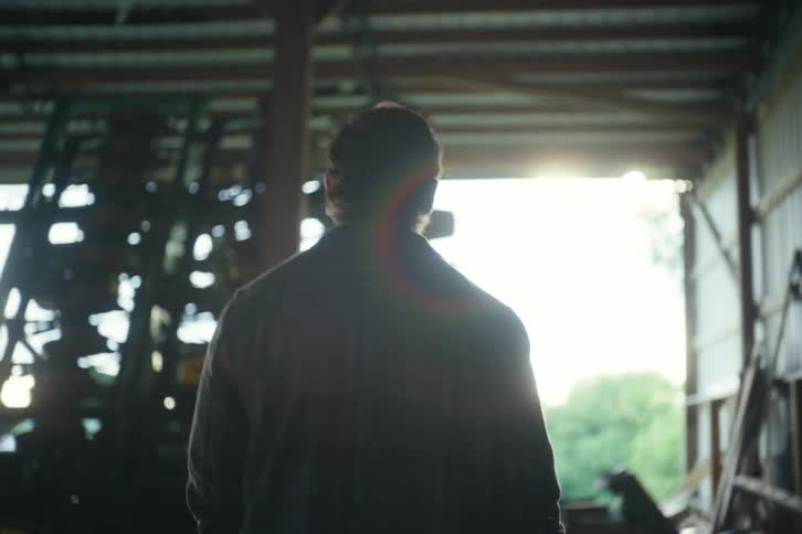 A silhouetted farmer walks in slow motion through a barn towards the bright sunlight at the entrance