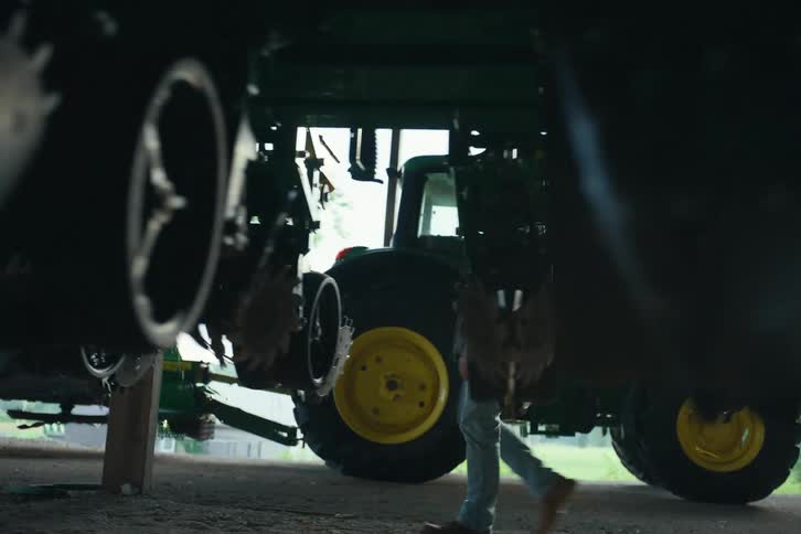 A farmer walks underneath large green farm equipment while performing an inspection inside of a barn