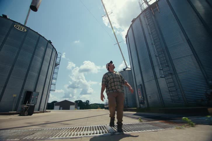 A farmer in a plaid shirt and hat walks confidently between towering metal grain silos on a sunny day