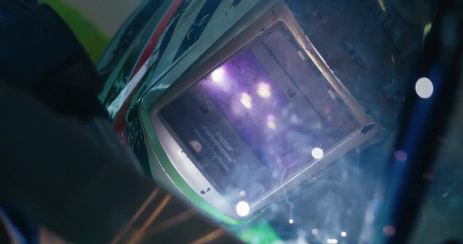 A close-up slow-motion shot captures a welder in a protective mask as sparks and smoke billow from the bright arc