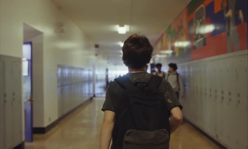 Slow motion shot from behind of a teenage boy with a backpack walking down a long, locker-lined high school hallway