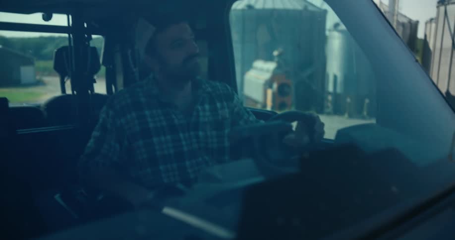 A farmer with a mustache wearing a plaid shirt drives a utility vehicle out from a shaded area into the sun