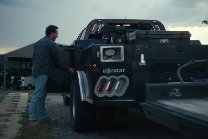 A man gets out of his large black work truck at dusk, retrieves a tool and a drink from a side compartment, and walks toward the camera