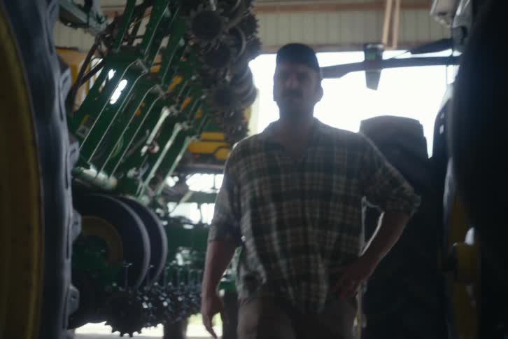 Low-angle slow-motion shot of a male farmer in a plaid shirt and hat walking past his large farming machinery in a barn