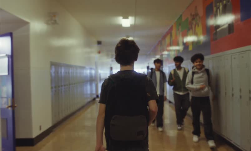 From a blurry background, a male student walks down a high school hallway lined with lockers in slow motion