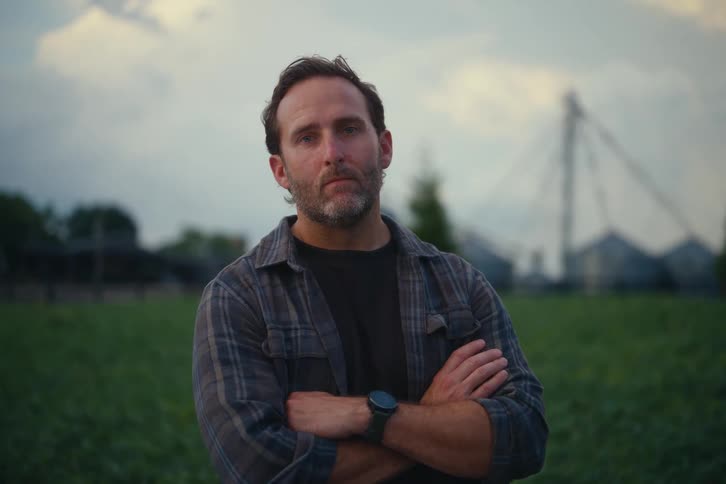 A serious farmer stands with his arms crossed in a field at dusk, looking directly at the camera