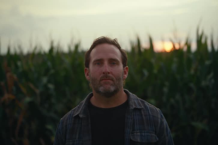 Portrait of a bearded farmer in a plaid shirt with a cornfield behind him at sunset