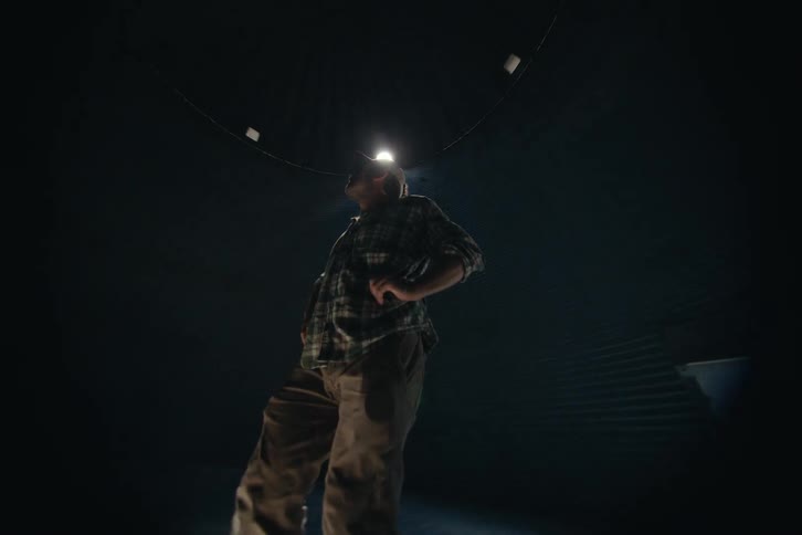 A farmer wearing a headlamp stands inside a dark and empty grain bin, inspecting the large structure