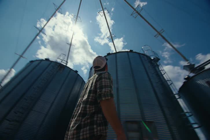 A low-angle tracking shot follows a farmer as he walks through a farmyard with large, imposing grain silos on a sunny day