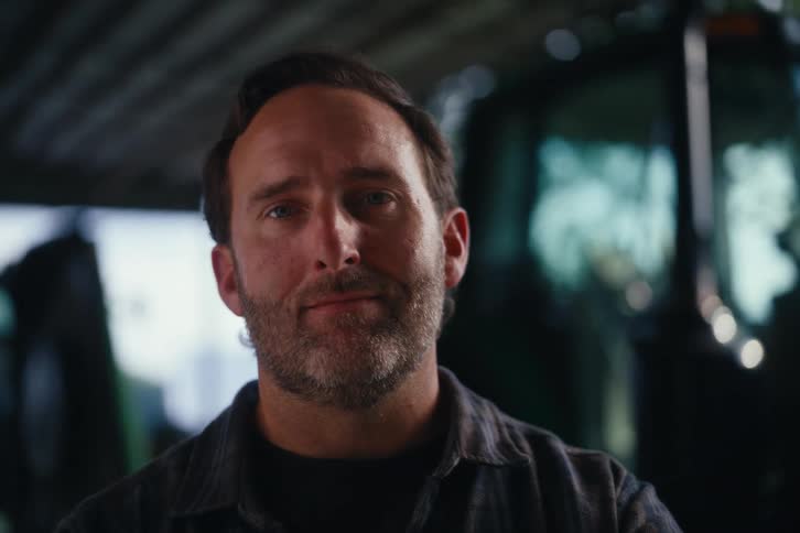 A pensive farmer with a beard turns to look at the camera with a subtle smile while standing in a barn in front of his tractor