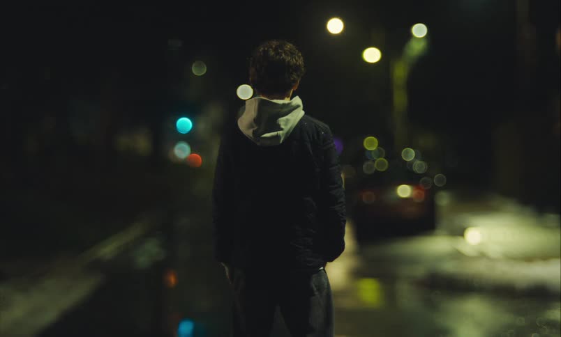 Rear view of a man standing on a snowy city street at night, with out-of-focus city lights creating a bokeh effect