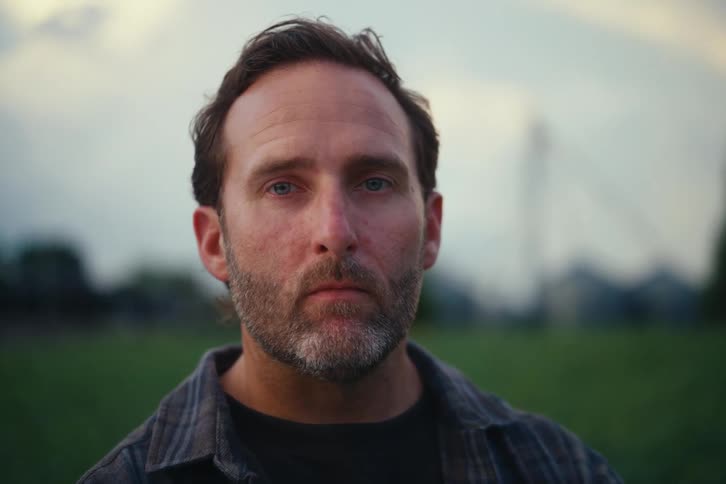 A close-up portrait of a serious, bearded, middle-aged man staring intently into the camera while standing outdoors at dusk