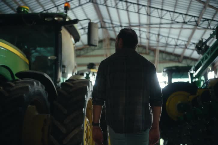 A farmer walks in slow motion through a large barn filled with his tractors and farming equipment